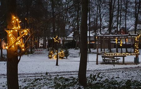 Verschneiter Stadtpark mit festlicher Beleuchtung in Bäumen und einem Pavillon, vorabendliche Stimmung.
